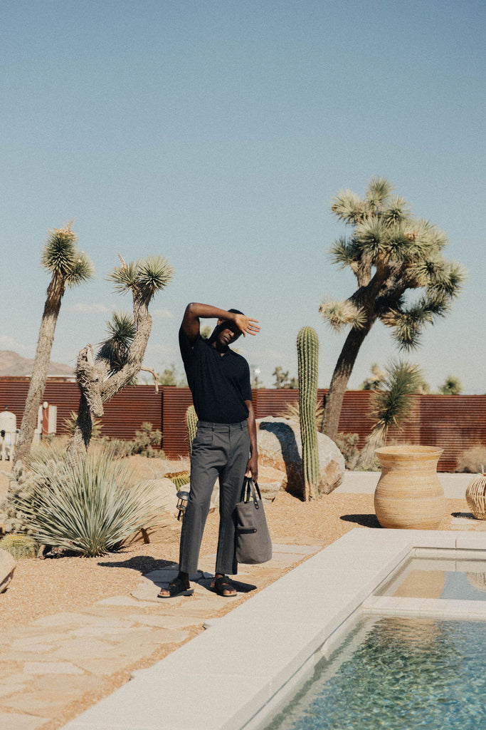 A man stands next to a pool shielding his eyes from the sun holding a leather-handled canvas tote