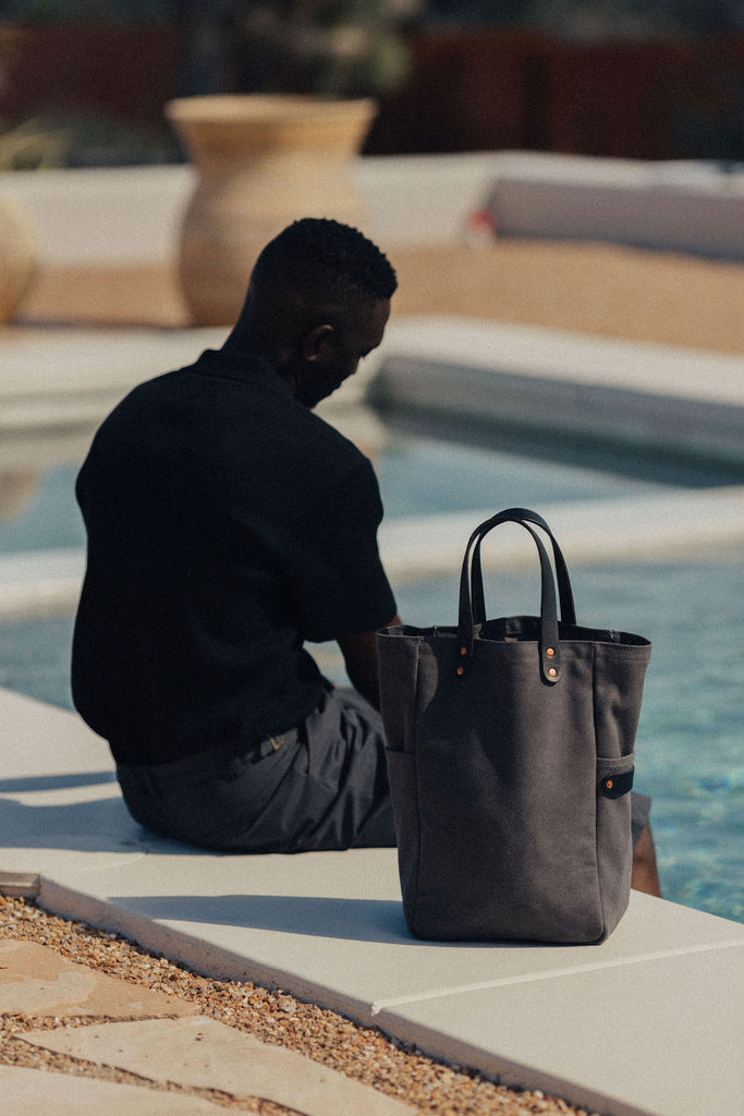 A man sits with his feet in a pool and has a leather-handled canvas tote next to him