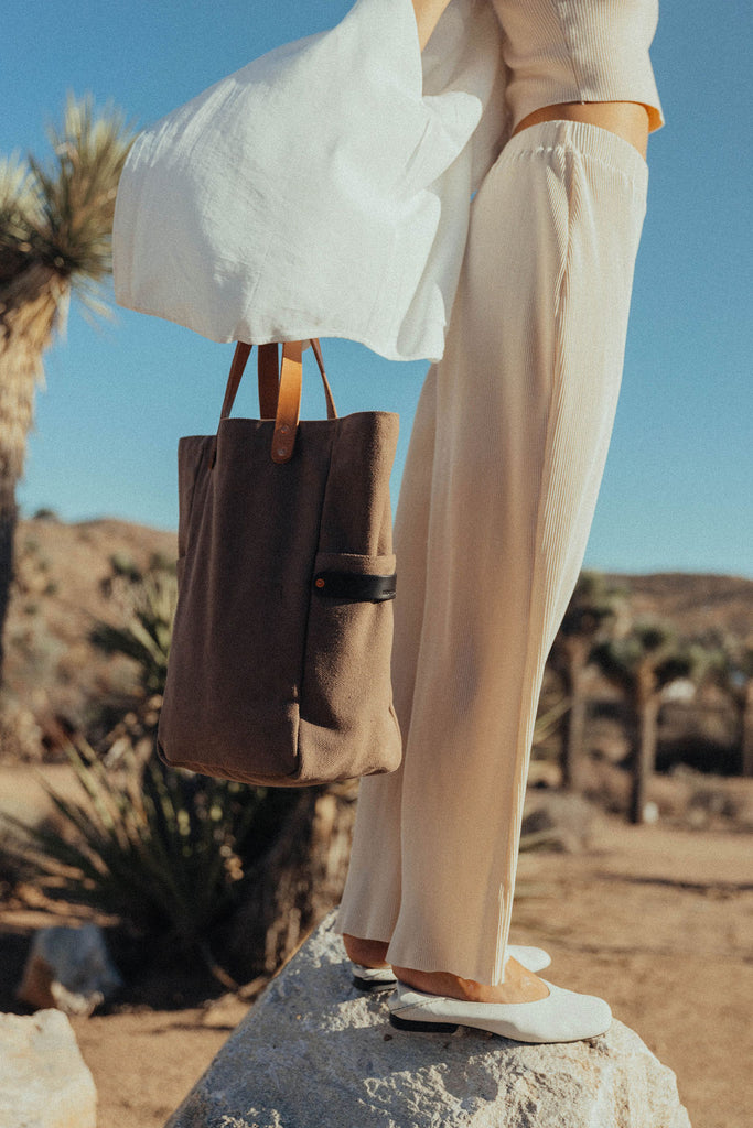 A woman standing on a boulder, the wind blowing her white cotton shirt holding a leather-handled canvas tote in her hand