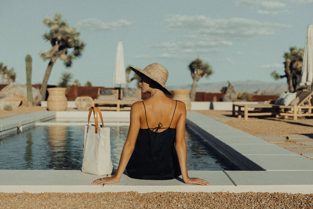 A woman in a sun hat sits with her feet in a pool and a leather-handled canvas tote next to her