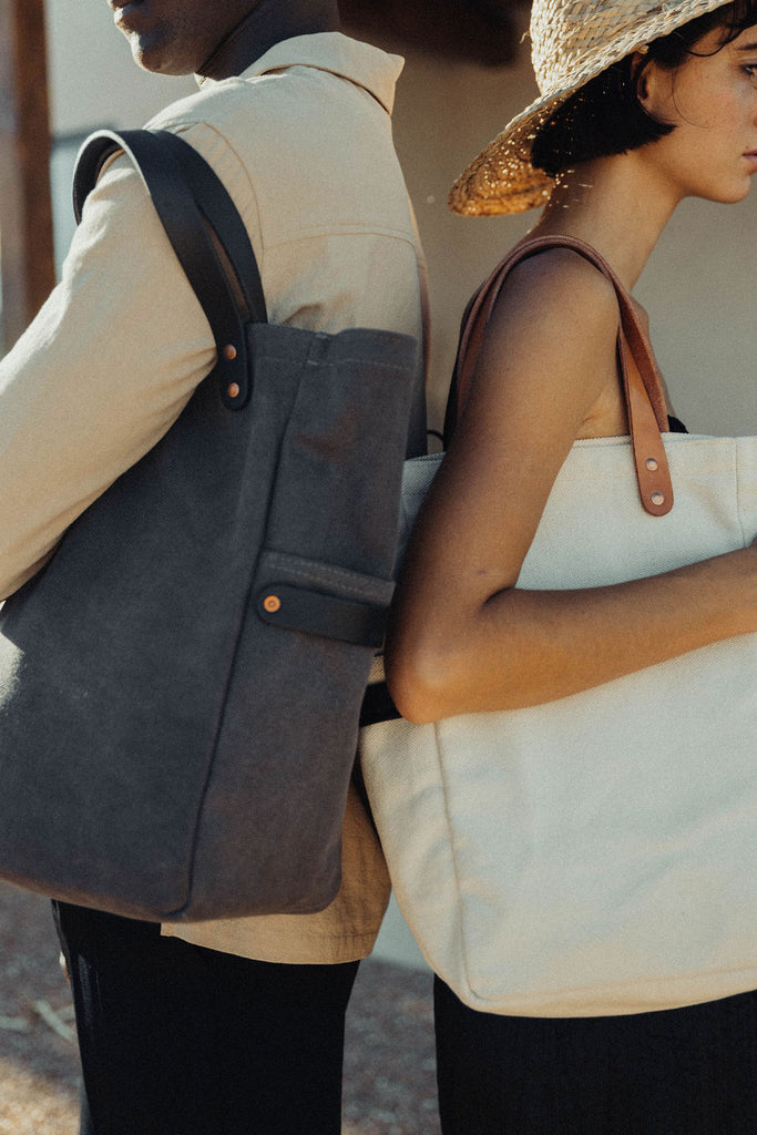 A man and woman stand back to back with leather-handled canvas totes on their shoulders