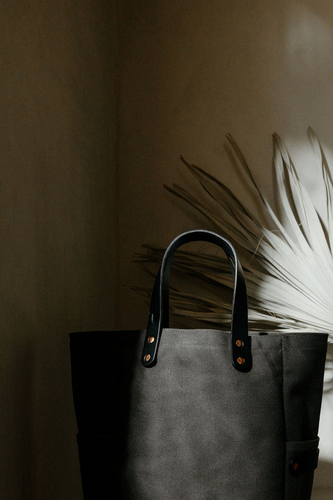 A leather-handled canvas tote in smoke with a dry palm branch behind it