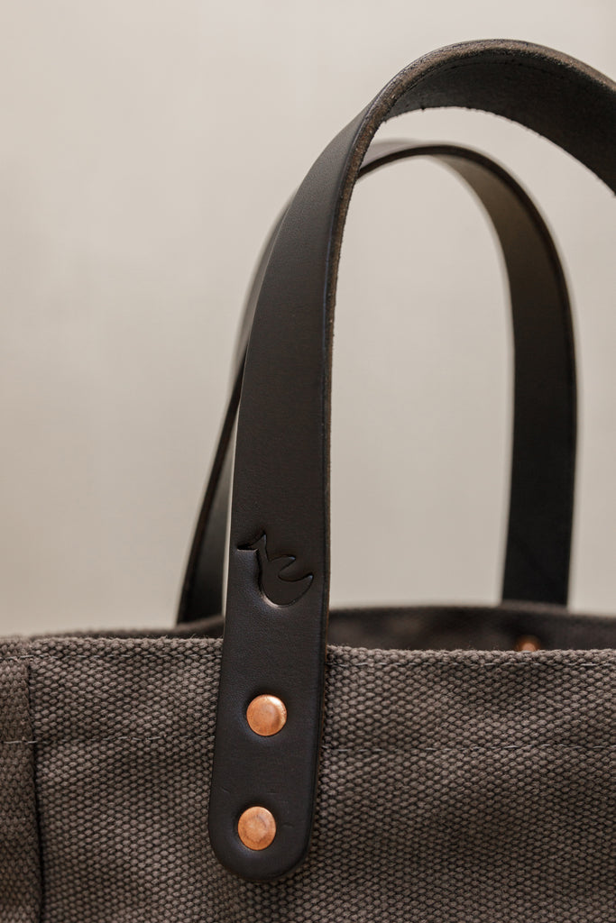 A closeup of leather handles with embossed bird and copper rivets on a leather-handled canvas tote in smoke